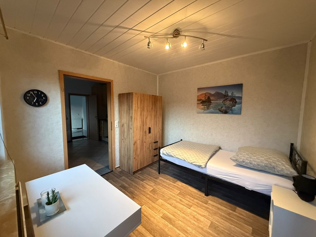 Zimmer mit Einzelbett, Holzkleiderschrank und weißem Tisch; offene Tür zum angrenzenden gefliesten Bereich.
